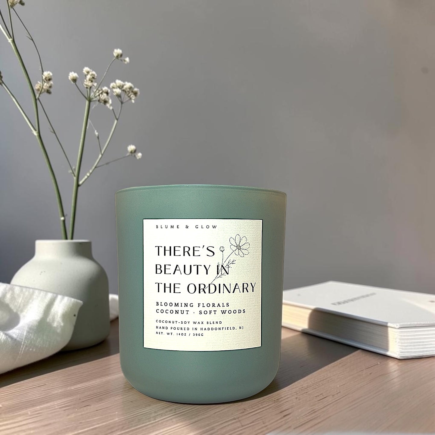 A matte sage green candle jar with a white label reading 'THERE'S BEAUTY IN THE ORDINARY' sits on a wooden table beside a white vase with dried stems and an open book.