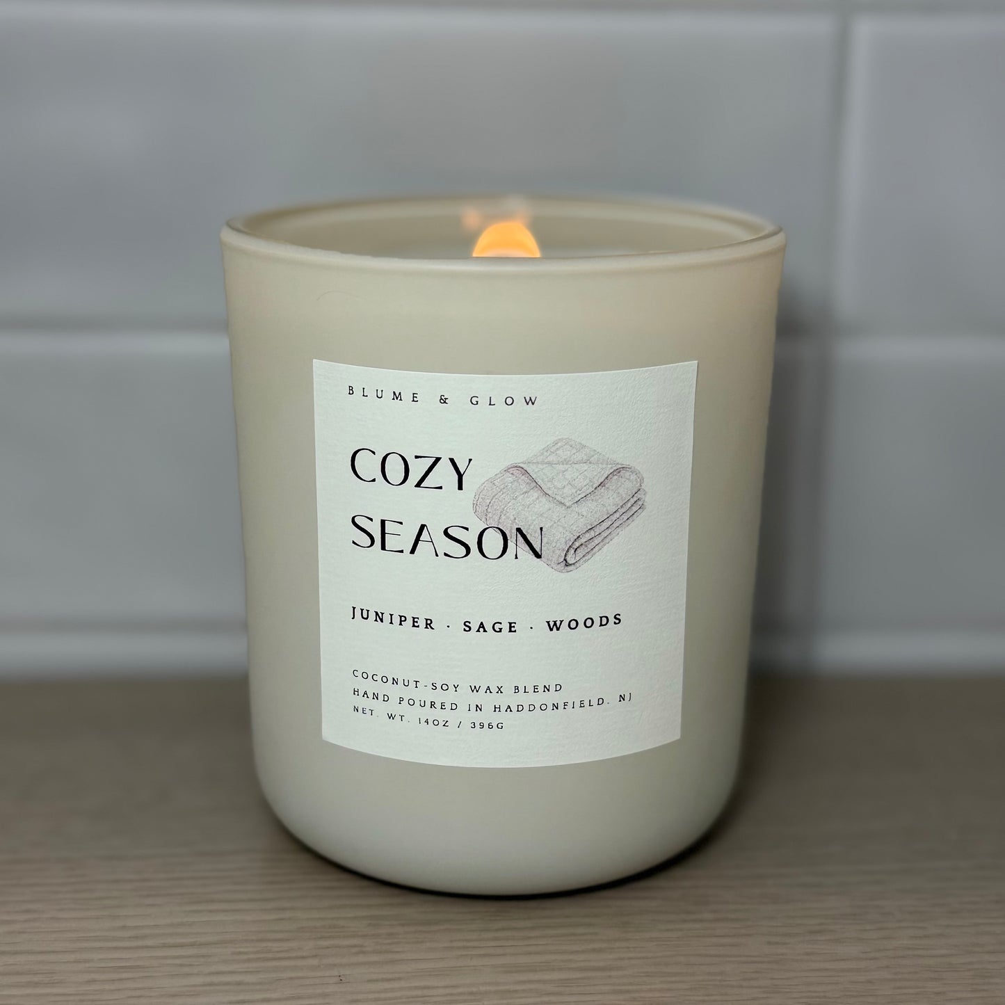 Cozy Season candle burning with a glowing wooden wick in a cream glass vessel.