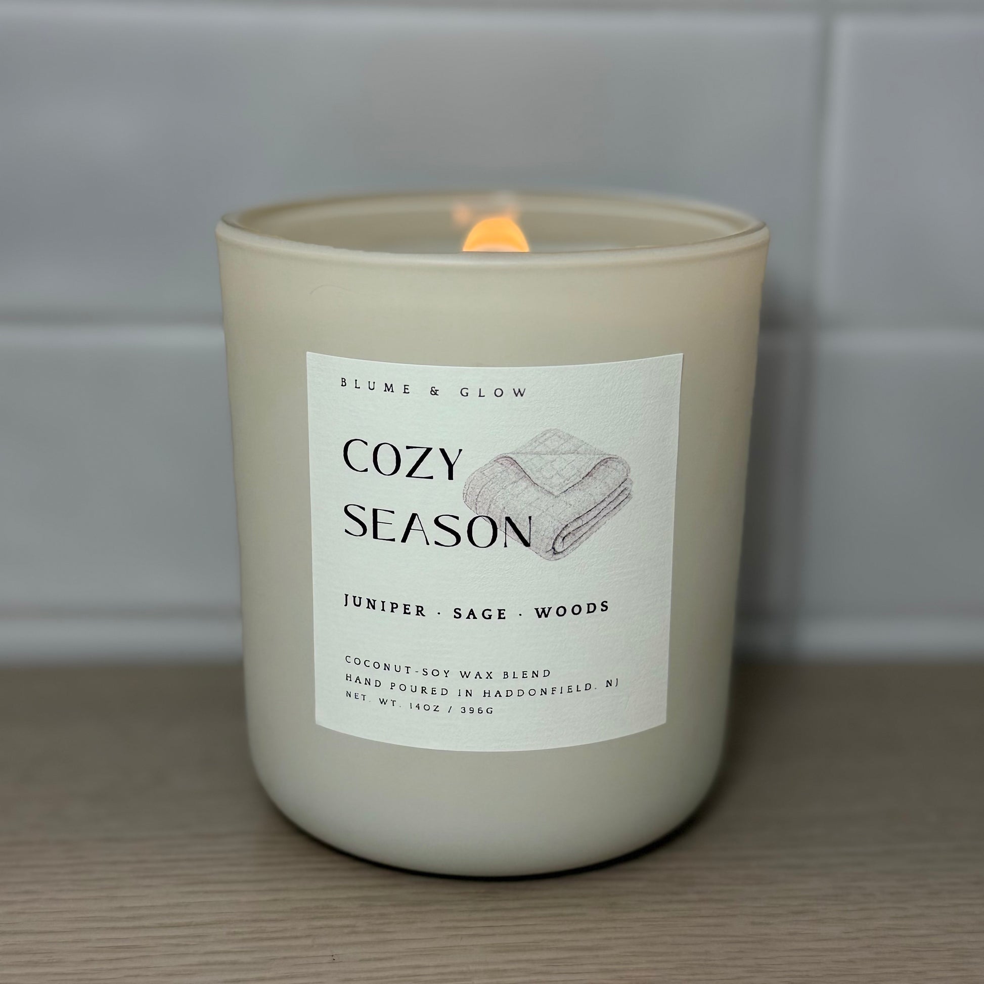 Cozy Season candle burning with a glowing wooden wick in a cream glass vessel.