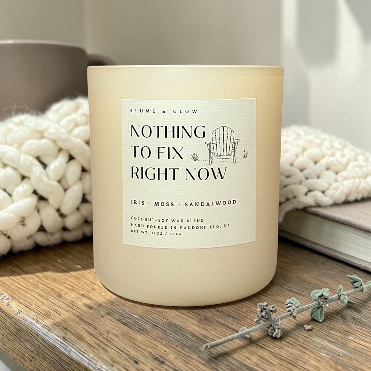 A matte cream-colored candle jar with a white label reading 'NOTHING TO FIX RIGHT NOW' by Blume & Glow, styled on a wooden surface with a chunky knit blanket, a book, and a dried sprig in the background.
