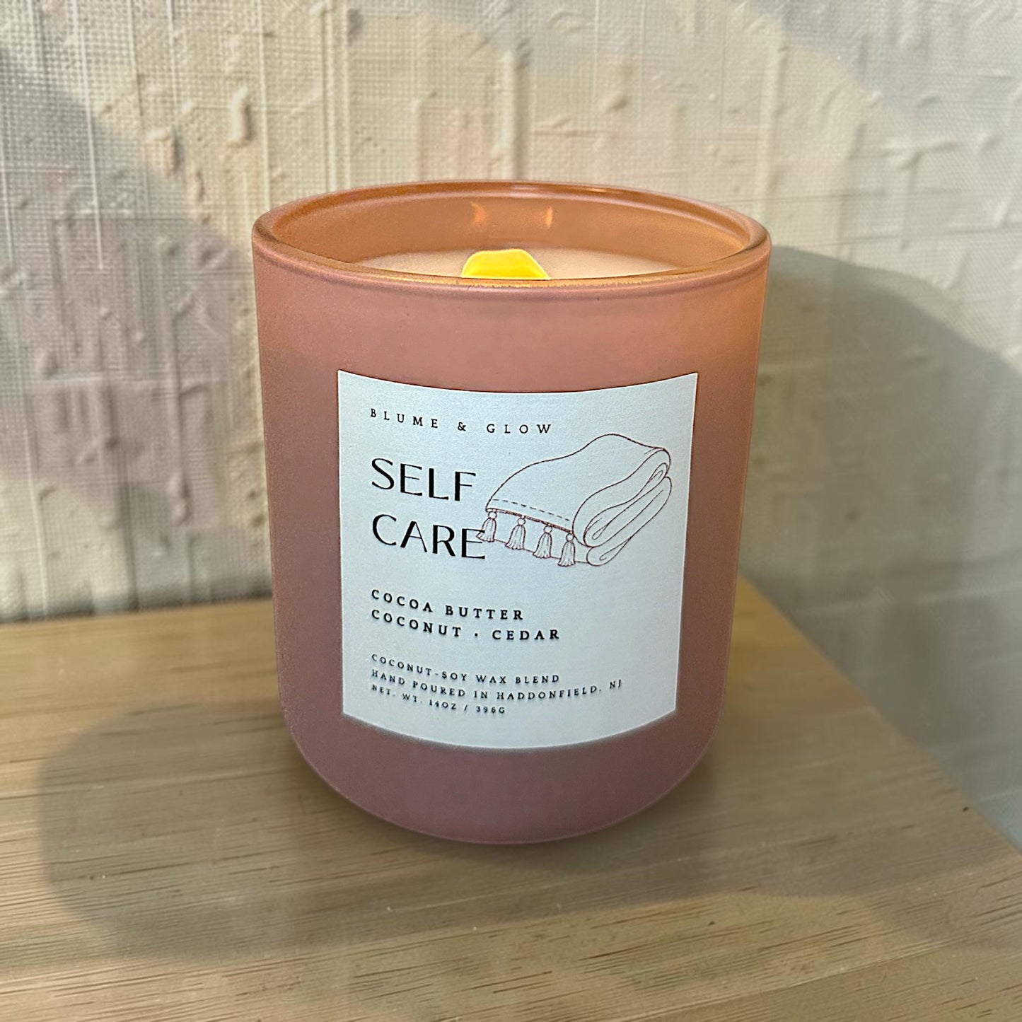 A lit scented candle in a matte blush jar with a white label reading 'SELF CARE' and 'Blume & Glow,' set on a light wood surface against a textured white wall.
